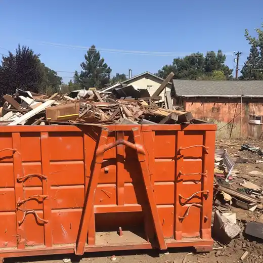 A large orange roll-off dumpster overflowing with construction debris, including scrap wood, roofing materials, and renovation waste. The container is located at a residential job site, showcasing high-capacity construction waste disposal and reliable debris hauling services for home remodeling projects.