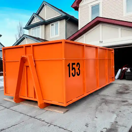 A large orange roll-off dumpster staged on a concrete residential driveway using protective wood planks to prevent ground damage. The container is positioned in front of a modern suburban home, highlighting professional driveway-friendly dumpster rentals for large home cleanouts and roofing projects.