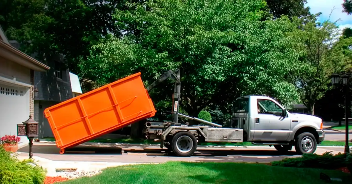 A professional roll-off dumpster delivery in progress, featuring a bright orange waste container being carefully lowered from a silver hook-lift truck onto a residential driveway. The scene is set in a quiet neighborhood with lush green trees and a house in the background, highlighting efficient waste management and debris removal services.