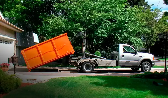 A professional roll-off dumpster delivery in progress, featuring a bright orange waste container being carefully lowered from a silver hook-lift truck onto a residential driveway. The scene is set in a quiet neighborhood with lush green trees and a house in the background, highlighting efficient waste management and debris removal services.