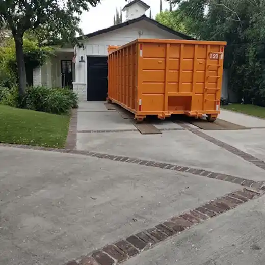 A large orange roll-off dumpster neatly placed on a residential concrete driveway, using protective wooden boards to prevent surface damage. The scene features a white suburban home with well-manicured landscaping, showcasing professional driveway-safe dumpster rental and residential waste disposal services.