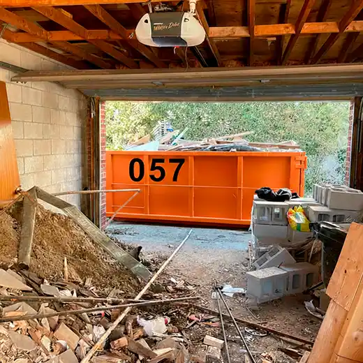A view from inside a messy garage looking out at a large orange roll-off dumpster filled with debris. The foreground shows piles of dirt, wood, and concrete blocks, illustrating a residential garage cleanout and on-site construction waste management solution.