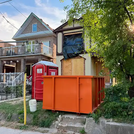 A small orange roll-off dumpster and a red portable toilet stationed in front of a residential home undergoing major renovations. The house features boarded-up windows and plastic coverings, illustrating comprehensive job site equipment rentals and residential demolition waste management services.